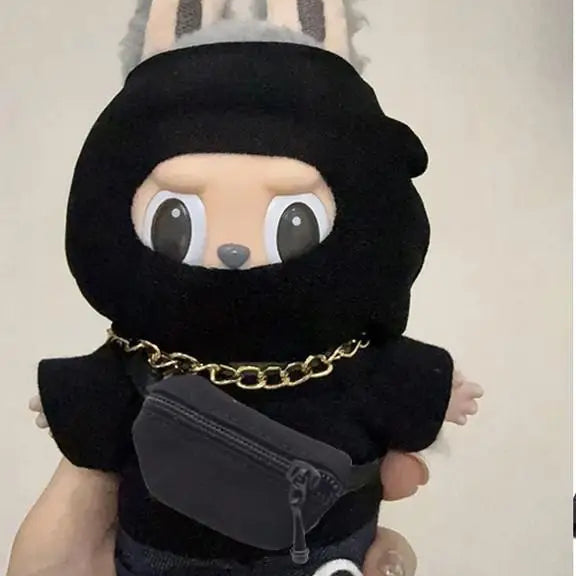 Plush toy dressed as a ninja with a black hood and outfit, held by a hand against a neutral background.